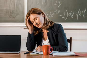 tired female teacher with eyes closed sitting at computer desk in classroom