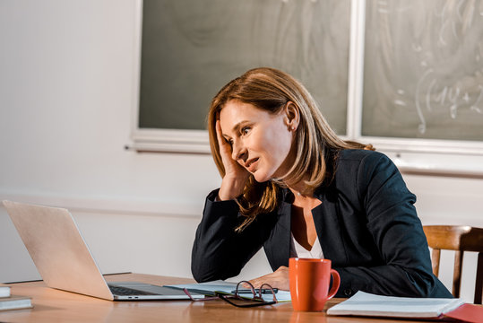 Tired Female Teacher Sitting At Computer Desk During Lesson In Classroom