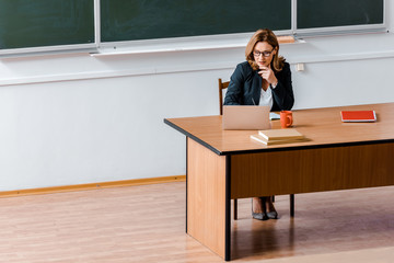 female university professor in glasses sitting at desk and using laptop in classroom
