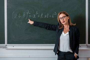 female teacher looking at camera and pointing with finger at mathematical equation on chalkboard in class