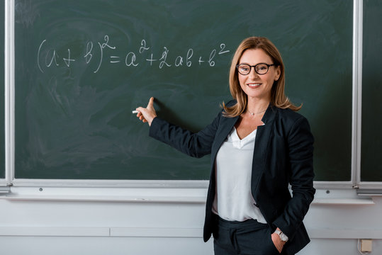 Female Teacher Pointing With Finger At Mathematical Equation On Chalkboard In Class