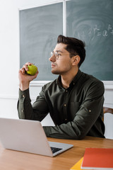 focused male teacher in glasses sitting at computer desk and holding apple in classroom