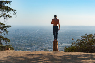 Man standing on a dead tree stump in Griffith park looking out over the city of Los Angeles though hazy sunlight