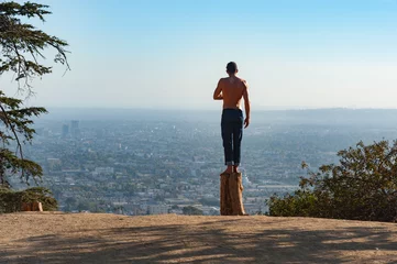 Tuinposter Man standing on a dead tree stump in Griffith park looking out over the city of Los Angeles though hazy sunlight © Gabriel Cassan
