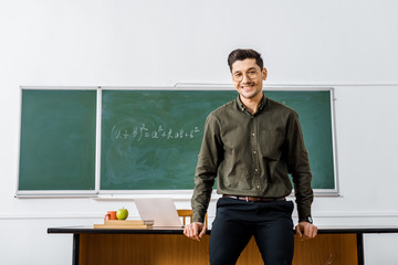 smiling male teacher in formal wear looking at camera and standing near desk in classroom