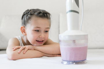 The child observes how the cocktail is made in the mixer. The girl looks like a blender mixes berries, fruit and milk.