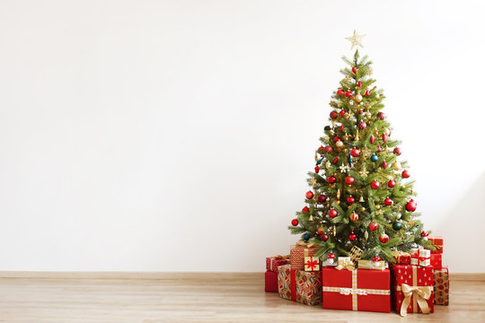 Big beautiful christmas tree decorated with beautiful shiny baubles and many different presents on wooden floor. White wall background with a lot of copy space for text. Close up.
