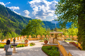 A woman admires the beautiful garden of Hohenschwangau castle with the Lion Fountain, the small Goose Man Fountain, flowers, trees and benches; surrounded by the forest and the Alps in the background. - obrazy, fototapety, plakaty