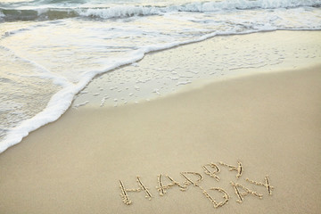handwritten inscription happy day made in the sand near the water, the wave washes text on the beach