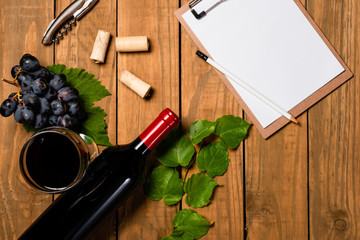 Composition with bottle and glass of wine with grape bunch and vine leaves and notepad on wooden background. Top view with copy space.