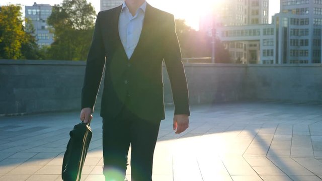 Young businessman with a briefcase walking in city street. Business man commuting to work in the urban environment. Confident guy in suit being on his way to work. Cityscape background. Slow motion