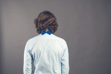 Back side view of a man with dark shaggy curly hair wearing blue shirt and bow tie against a grey background. Studio shot. Rear view people collection. Back view of person. Toned. Space for text.