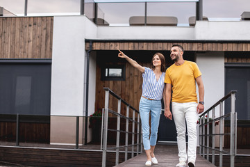 Full length portrait of happy lady holding hands with her boyfriend and pointing aside. They looking there with smile. Copy space on left side