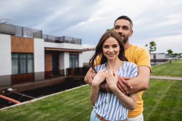 Waist up portrait of cheerful man hugging his girlfriend while standing in front of house. She looking at camera with smile. Copy space on left side