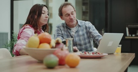 While taking the breakfast and drinking coffee mature couple in the kitchen analyzing something from the laptop in the morning. 4k - Powered by Adobe