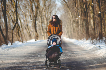 Young beautiful woman with stroller walking in the snowy park on sunny day in winter