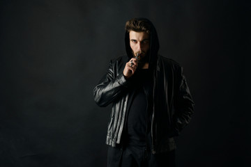 Man with vaping mod exhaling steam at black studio background. Bearded guy smoking e-cigarette to...