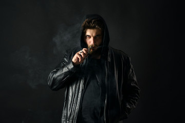 Man with vaping mod exhaling steam at black studio background. Bearded guy smoking e-cigarette to...