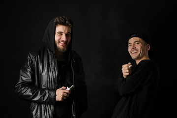 Bad habit, smoking in a public place. Two Young man blowing smoke to join it in one cloud at black studio background. Friends and vape addiction concept. 