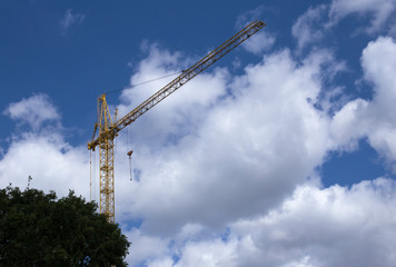 Construction crane against the blue sky and clouds. In the left corner are trees.