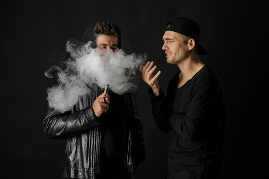 Bad habit, smoking in a public place. Two Young man blowing smoke to join it in one cloud at black studio background. Friends and vape addiction concept. 