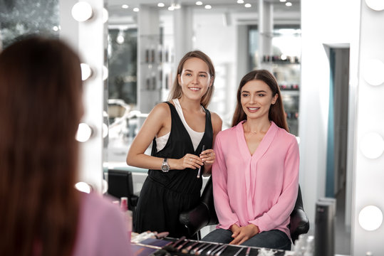 Waist up photo of professional make-up artist and pretty girl considering makeup while looking in the mirror