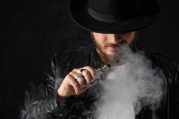 Man with vaping mod exhaling steam at black studio background. Bearded guy smoking e-cigarette to...