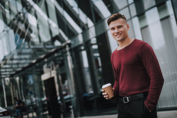 Concept of little break in work. Waist up side on portrait of successful smiling man staying with cup of coffee near business center. Copy space on left