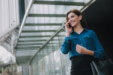 Concept of success and well-being. Waist up low angle portrait of smiling successful lady in stylish cloth talking by smartphone near business center. Copy space on left