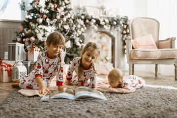 Two little sisters and a tiny brother lie on the carpet and read book near the New Year's tree with gifts in the light cozy room with armchair and fireplace