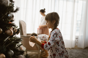 One little girl in pajama decorates a New Year's tree in the light cozy room