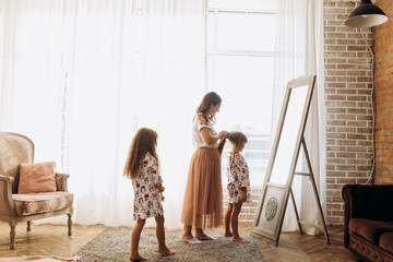 Young mother combing her little daughter's hair standing in front of the mirror and her second  daughter comes to them in the full of light cozy room