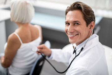 Cheerful emotional young doctor smiling happily while sitting behind his patient with stethoscope