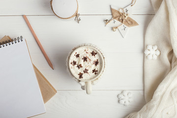 A cup of hot winter drink, with whipped cream and powder with an asterisk, a notebook, white snowflakes and a knitted scarf on a wooden table. Christmas concept in bright colors. Copy space.