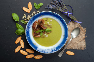 Soup puree from young green peas with bacon, pumpkin seeds and basil leaves in a plate with a blue border. Delicious and healthy lunch.