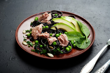 Pasta with cuttlefish ink (black noodles) with tuna pieces, basil leaves, avocado slices, green peas, onions and sunflower seeds on a clay plate. An original pasta recipe. Useful and tasty dish