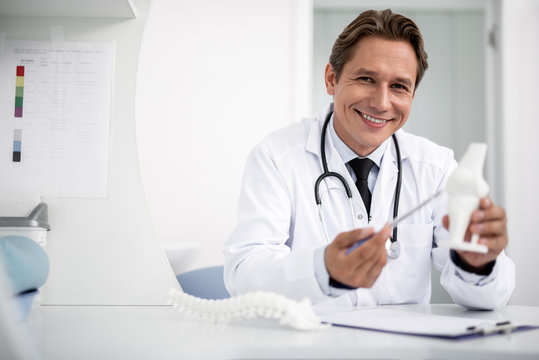 Emotional cheerful doctor in white uniform kindly smiling and demonstrating the knee model