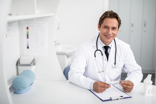 Cheerful friendly practitioner sitting with his hands on the table and smiling while having stethoscope on his neck