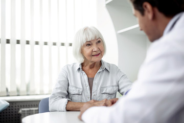 Calm elderly lady sitting in front of her practitioner and feeling interested while talking to him and smiling