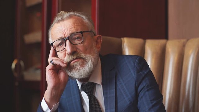 Serious pensive old male in formal blue suit, keeps palms near his chin, thinking about his children while waiting them at home.