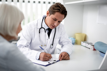 Attentive professional practitioner writing on the clipboard while sitting at the table with patient next to him