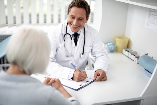 Positive emotional general practitioner sitting with the clipboard on the table and kindly smiling to his patient