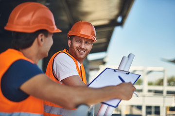 Positive friendly builder in orange helmet looking at his coworker and smiling while standing next...