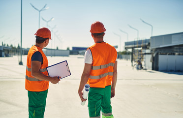Two professional builders in orange vests and helmets standing with drawings and notes while...