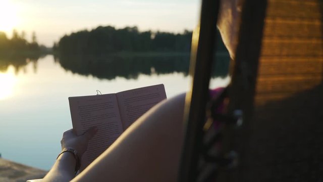Young white millenial woman with glasses reading book while sitting in chair on pier overlooking beautiful scenic lake vista with trees at sunset