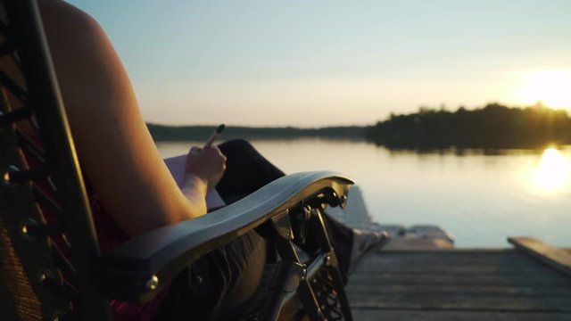 Young white millenial woman journaling while sitting on pier overlooking sunset at beautiful scenic lake