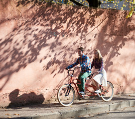 Fototapeta na wymiar Young attractive tourist couple in casual clothing, bearded man and blond woman in glasses cycling tandem bike along empty paved sunny sidewalk on background of high red crackled plastered wall.