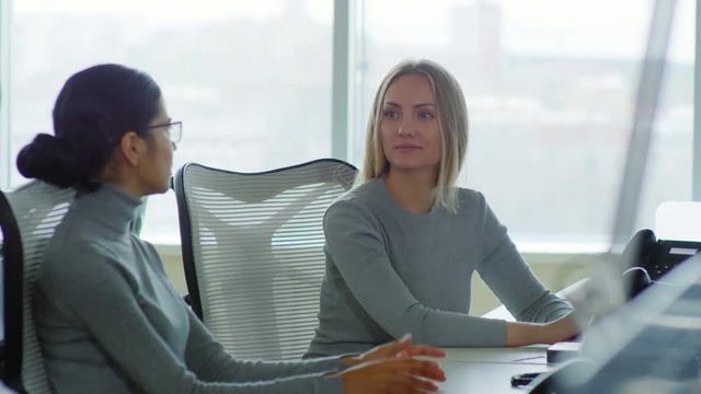 Medium shot of two young women sitting at desk in office and talking during break from work