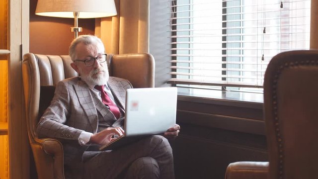 Fashionable focused elderly male writer wearing glasses and elegant clothes working on laptop, close up. Old author creating new story while sitting in big leather arm chair near the window.