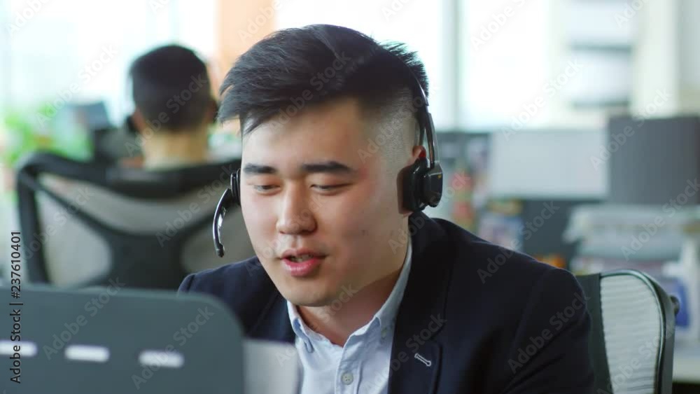 Poster medium shot of young asian man wearing headset and using desktop computer when talking to client